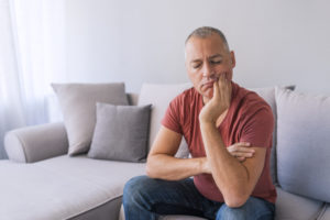 Photo of Depressed ill man having toothache and touching cheek. Mature man suffering from tooth pain, caries. Handsome gray hair male suffering from toothache, closeup. Portrait of casual 46 years mature man toothache with painful expression, sitting on sofa at home,