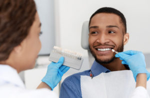Female dentist choosing filling shade for smiling black guy, using tooth scale sample, close up