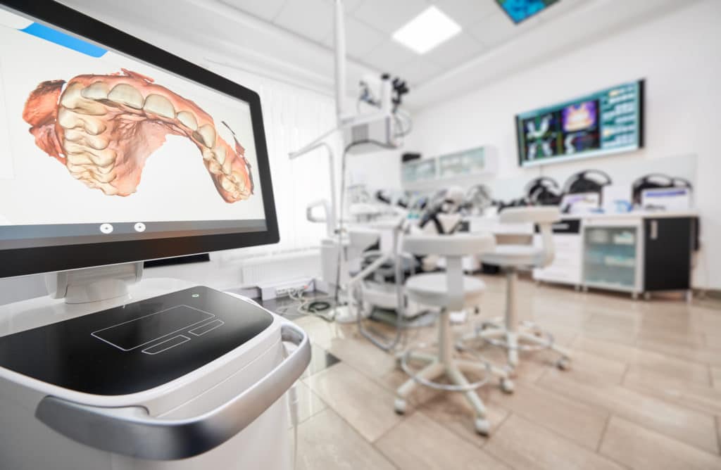 close up of computer screen in dental office displaying model of teeth with various dental equipment in room in center and right
