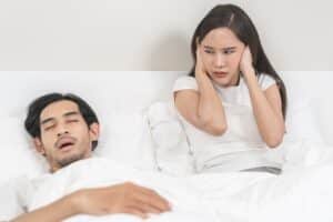 Woman plugging her hears as man snores heavily while sleeping in bed.
