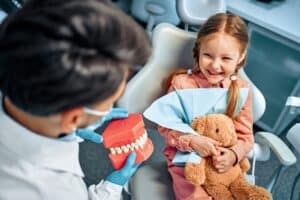 Young girl smiling in a dental chair holding a stuffed animal while a dentist shows her a model of teeth.